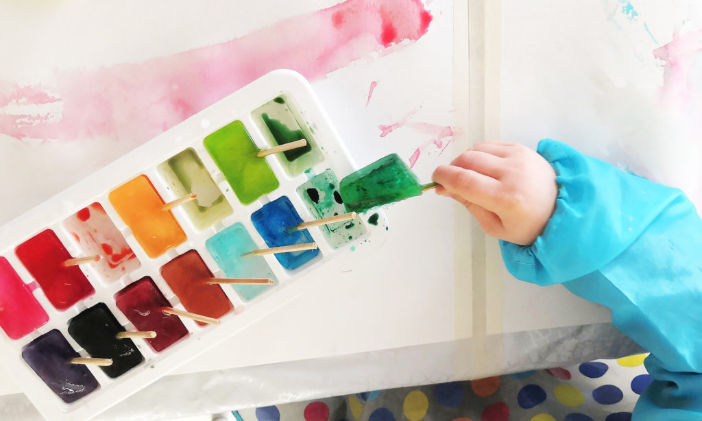 Kuvassa erivärisiä jääkuutiopaloja muotissa, joilla lapsi maalaa paperia.