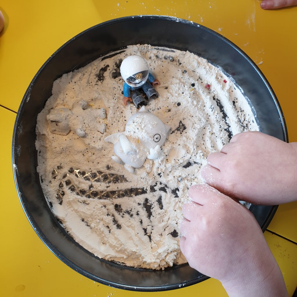 Kuvassa leivontavuoka johon taputeltu tasaiseksi pohjaksi jauhoja. Päälle ripoteltu pippuria, strösseliä ja siemeniä. Lapsi leikkii legohahmolla "kuussa".
