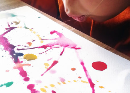Kuvassa lapsi puhaltaa paperille tiputettuja väripisaroita rikki.