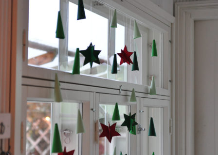 Jouluinen ikkuna, jossa roikkuvat siimalla itse askarrellut paperiset joulutähdet ja -kuuset.