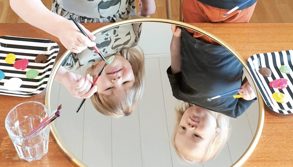 Kaksi lasta maalaa peiliin. Kuvassa lasten kasvot näkyvät peilin kautta.