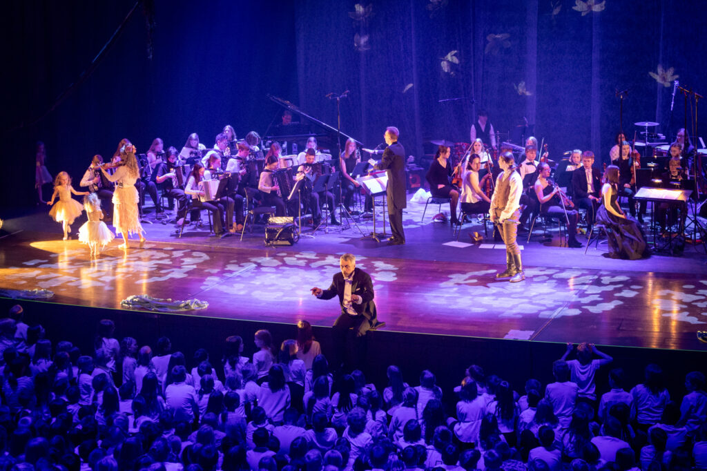 kuoronjohtaja johtaa suurta lapsikuoroa, lavalla tanssijoita, nuori Sibelius ja orkesterit, sininen valo