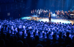 kuoronjohtaja johtaa suurta lapsikuoroa orkesterin säestäessä, sininen valo